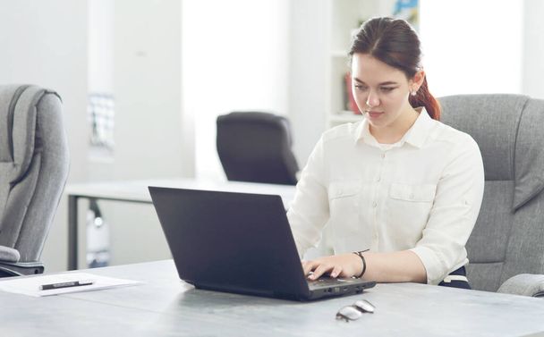 Молода приваблива бізнес-леді працює в офісі посміхаючись дивлячись в ноутбук
.  - Фото, зображення