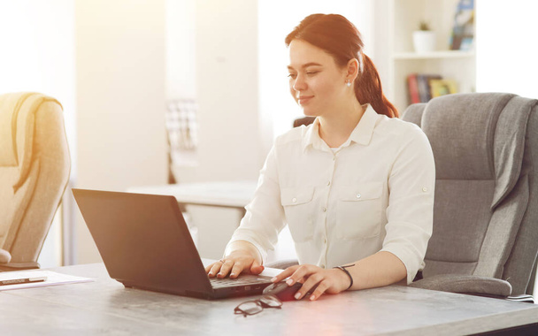 Jonge aantrekkelijke zakenvrouw werken in kantoor glimlachen op zoek naar laptop.  - Foto, afbeelding