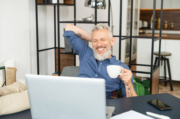 Cheerful mature male employee enjoys morning coffee at the workplace in the office - Foto, immagini