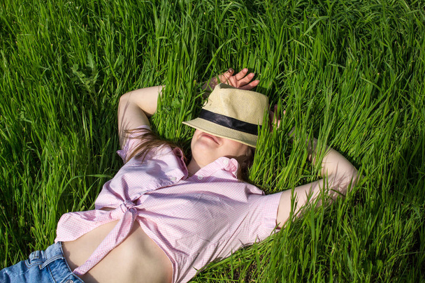 On a sunny day, a woman in a pink shirt lies on the green grass and hides her face with a straw hat Concept life style ecology colorful happy people freedom wonderfully beautiful, greens, environment - Photo, Image