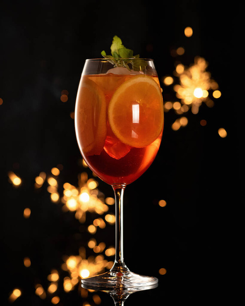 Lasi eksoottisia cocktail jään ja appelsiinin viipaleita mustalla pohjalla bokeh alkaen sparklers. Alkoholijuomaa. Pystysuora muoto - Valokuva, kuva