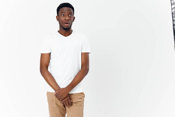 скромний африканець у білій футболці і бежевих штанях тримає руки перед собою на світлому фоні. - Фото, зображення