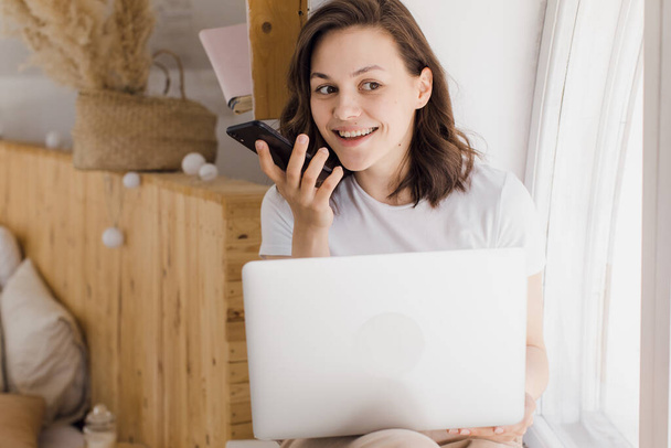 Smiling young Caucasian woman record digital audio message on smartphone in modern flat - Φωτογραφία, εικόνα