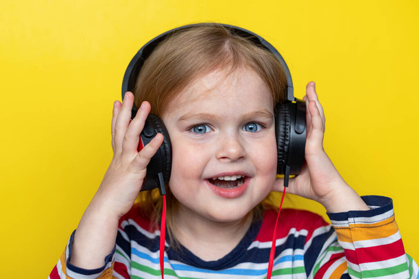 Szczęśliwe uśmiechnięte dziecko słuchające muzyki z dużymi słuchawkami, odizolowane na żółtym tle. - Zdjęcie, obraz