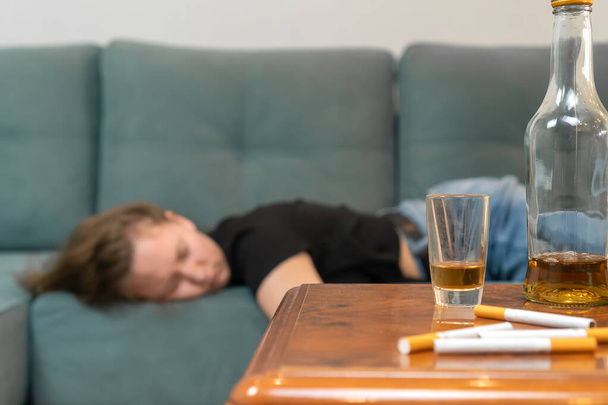 Пьяная молодая темноволосая женщина спит на диване после вечеринки, пустые бутылки на столе. Фокус на бутылках - Фото, изображение