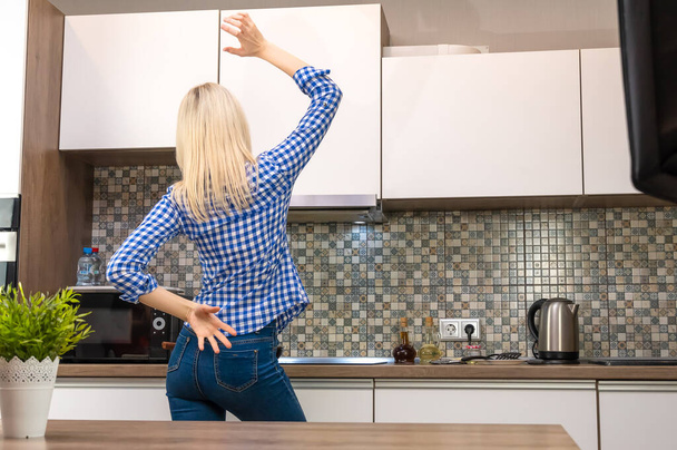 Cheerful young slender blonde woman in blue jeans and a shirt cooks in the kitchen and dances, copy space. Photo from the back - 写真・画像