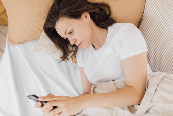 a young attractive girl is lying in bed and checking social networks. Modern bright apartment - Foto, imagen