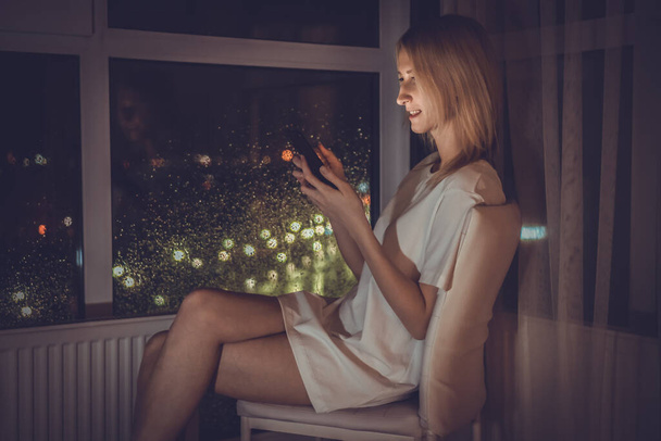 Hermosa mujer rubia joven con una camisa blanca con un teléfono móvil en las manos está sentada en una silla cerca de la ventana del piso al techo y la ciudad de la noche, la luz cálida del teléfono cae en su cara - Foto, Imagen