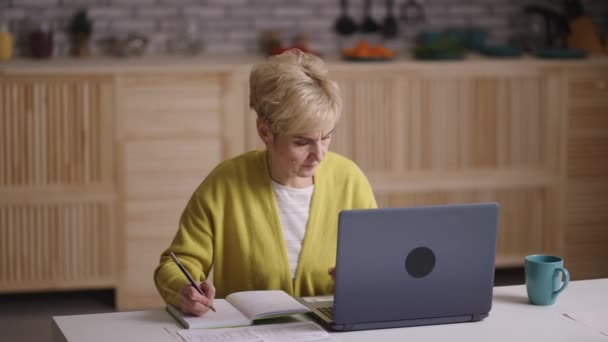samovýchova pro důchodce, starší žena prohlíží výuku na internetu pomocí notebooku a psaní poznámek, sedí doma - Záběry, video