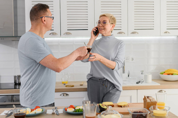 Mature couple enjoying coffee breakfast together at kitchen. Woman talking at phone - Photo, Image