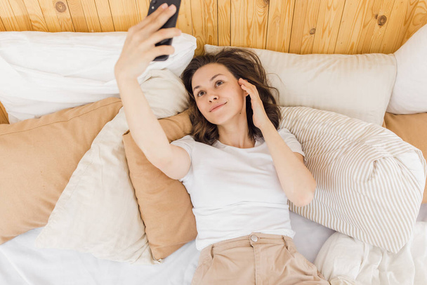 Close-up of an excited woman laughing, holding a smartphone, taking a selfie, having fun with a gadget, lying on the bed, enjoying her leisure time - Fotó, kép