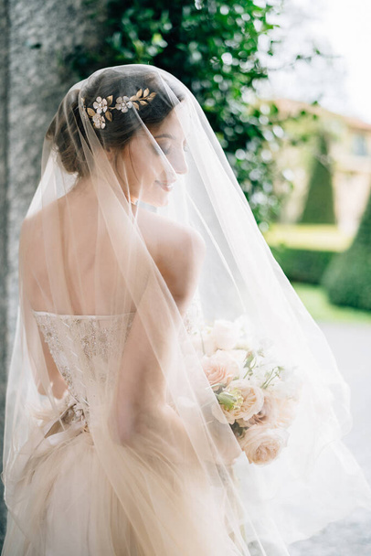 Novia en un vestido de novia con un velo y un ramo de flores está de pie con la cabeza vuelta cerca de la pared. Lago Como. Vista trasera. De cerca. - Foto, Imagen