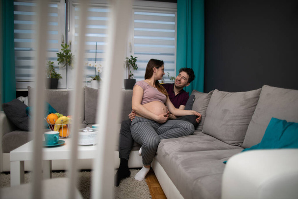 A husband hugs his pregnant wife. A married couple is sitting on a sofa in the living room at home. The pregnant woman. - Foto, imagen