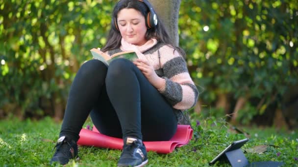 Νεαρή κοπέλα που διαβάζει ένα βιβλίο κάτω από ένα δέντρο σε έξαρση πανδημίας - Πλάνα, βίντεο