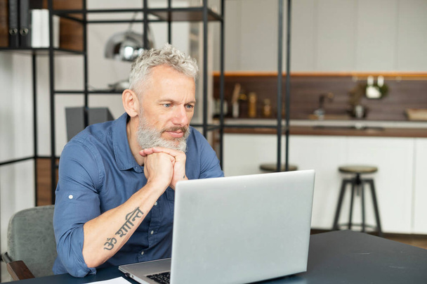 Serious middle age man in smart casual shirt lost in thoughts sitting at the desk with a laptop - Foto, Imagen