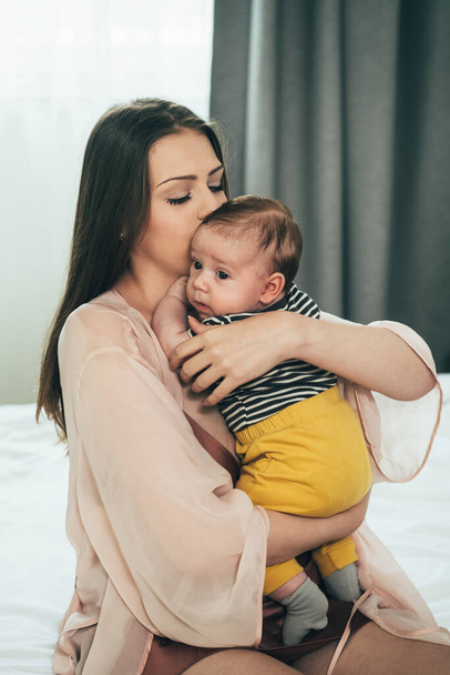 Young Mother Kissing and Holding her Baby in the BedroomCaring mother holding newborn baby in hands, embracing and kissing baby boy and enjoy together at home. - Photo, Image