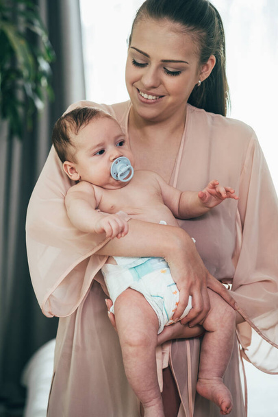 Πορτρέτο της χαμογελαστή μητέρα κρατώντας χαριτωμένο αγοράκι της σε Diaper .Happy νεαρή μητέρα στέκεται και κρατώντας νεογέννητο μωρό με πιπίλα στο στόμα. - Φωτογραφία, εικόνα