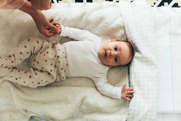 Retrato de bebé niño acostado en cuna y mirando a la camera.Cute bebé recién nacido sosteniendo la mano de la madre y descansando en la cama del bebé. - Foto, Imagen