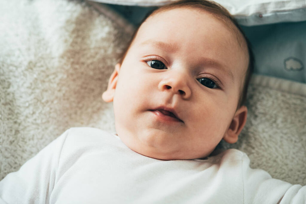 Κοντινό πλάνο Φωτογραφία του Αξιολάτρευτο μωρό Κοιτάζοντας CameraΠρόσωπο του περίεργου νεογέννητου αγοριού κοιτάζοντας την κάμερα και ξαπλωμένος στο κρεβάτι στο σπίτι. - Φωτογραφία, εικόνα