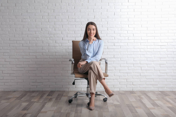 Young businesswoman sitting in office chair near white brick wall indoors - Foto, imagen