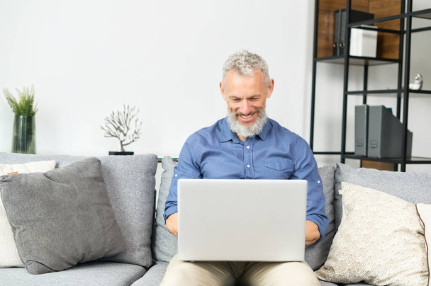 Modern middle-aged bearded man using laptop computer sitting on the couch at home - Zdjęcie, obraz