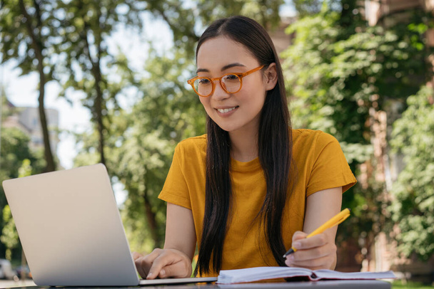 Όμορφη χαμογελαστή γυναίκα φορώντας κομψά γυαλιά χρησιμοποιώντας φορητό υπολογιστή, κρατώντας σημειώσεις, σχεδιάζοντας start up project, δουλεύοντας σε εξωτερικούς χώρους. Ασιατικές σπουδές, προετοιμασία εξετάσεων, online εκπαίδευση - Φωτογραφία, εικόνα