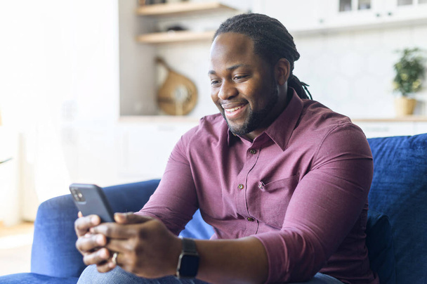 Smiling African-American man using smartphone sitting on the sofa at home - Valokuva, kuva