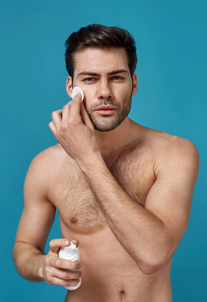 Sexy brunette naked man doing morning routine, cleansing his face with cotton pads before shaving and holding foam cleanser, posing isolated over blue background - Foto, afbeelding