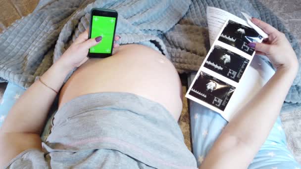 A pregnant woman is lying in bed with a phone in her hand holding her stomach, covering her legs with blanket, top view - Footage, Video