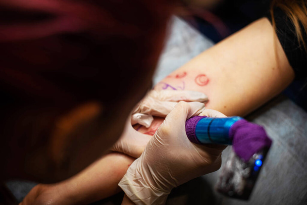 Bájos lány rózsaszín hajjal és tetoválásokkal egy tetoválást tesz egy nő kezére egy tetoválószalonban, kreatív kisvállalkozással és tetoválásmesterrel. Hipster nő kesztyűben rajzol egy tetoválást - Fotó, kép
