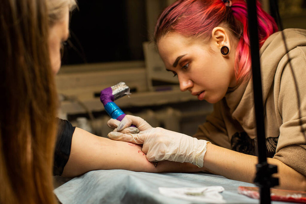 Urocza dziewczyna z różowymi włosami i tatuażami wypycha tatuaż na ręce kobiety w salonie tatuażu, kreatywnym małym biznesie i mistrzem tatuażu. Hipsterka w rękawiczkach rysuje tatuaż - Zdjęcie, obraz