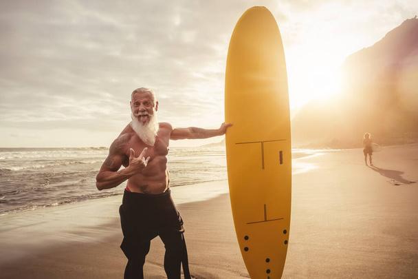Adatta a uomini anziani che si divertono a fare surf durante il tramonto - Addestramento dell'uomo in pensione con la tavola da surf sulla spiaggia - Stile di vita delle persone anziane sane e concetto di sport estremo - Foto, immagini