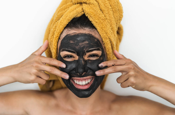 Jeune femme souriante appliquant un masque de charbon sur le visage - Bonne fille ayant soin de la peau spa jour à la maison - Beauté saine traitement propre et concept de style de vie des jeunes - Photo, image