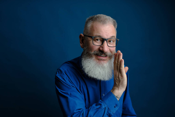 Bearded middle age man dressed blue shirt holds hand near mouth, secret talk concept, blue backdrop - Foto, Bild