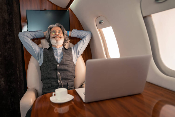Szczęśliwy biznesmen siedzi w pobliżu zamazanego laptopa i kawy w prywatnym samolocie  - Zdjęcie, obraz