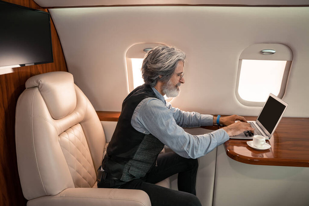Вид сбоку зрелого бизнесмена, использующего ноутбук возле чашки кофе в частном самолете  - Фото, изображение