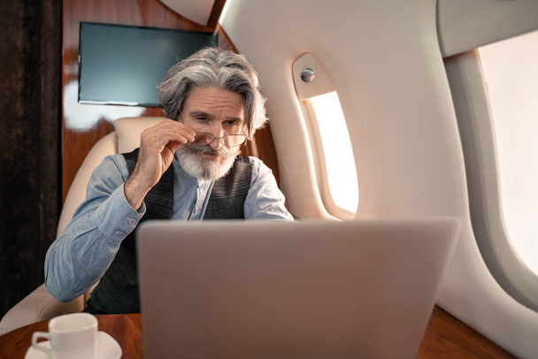 Érett üzletember kezében szemüveg közelében laptop és kávé privát jet  - Fotó, kép