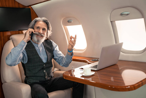 Hombre de negocios sonriente hablando en el teléfono inteligente cerca de la computadora portátil y café en el avión  - Foto, Imagen
