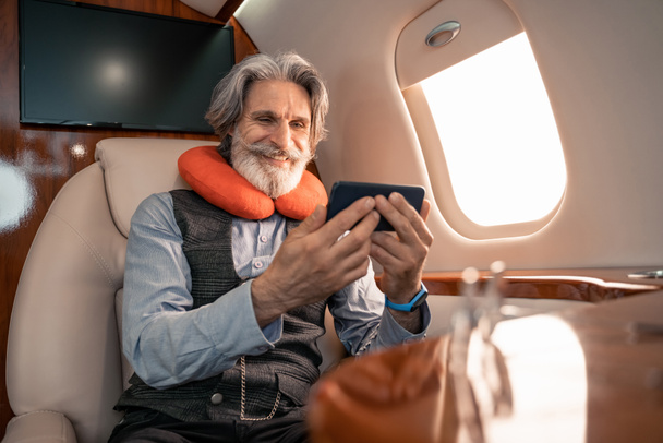 Uomo d'affari sorridente in cuscino collo utilizzando smartphone in aereo  - Foto, immagini