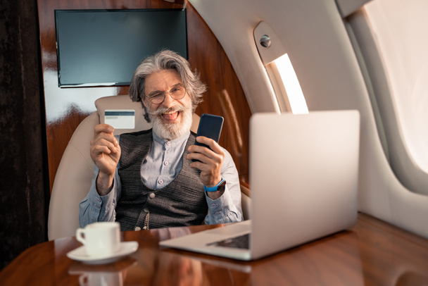 Бизнесмен с кредитной картой и смартфоном подмигивает возле ноутбука и кофе в струе  - Фото, изображение