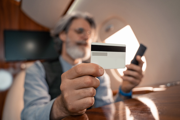 Credit card in hand of businessman using smartphone on blurred background in jet  - Photo, Image