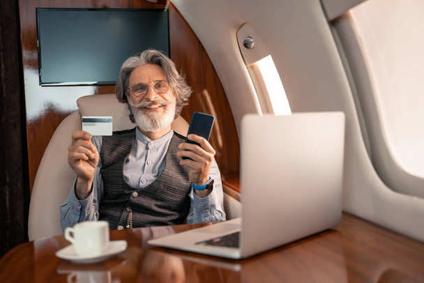 Gülümseyen işadamı özel uçakta dizüstü bilgisayar ve kahve yanında kredi kartı ve akıllı telefon tutuyor.  - Fotoğraf, Görsel