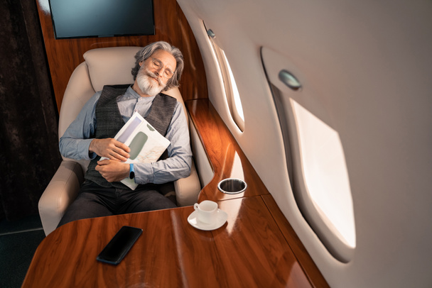 Durmiendo empresario celebración revista cerca de teléfono inteligente y café en avión privado  - Foto, Imagen