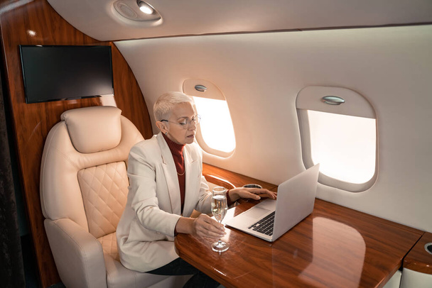 Mature businesswoman using laptop and holding glass of champagne in private plane  - Photo, Image