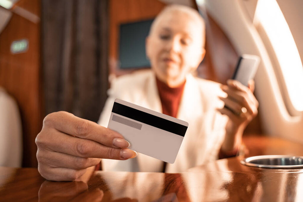 Kreditkarte in der Hand einer Geschäftsfrau mit Smartphone vor verschwommenem Hintergrund im Flugzeug  - Foto, Bild