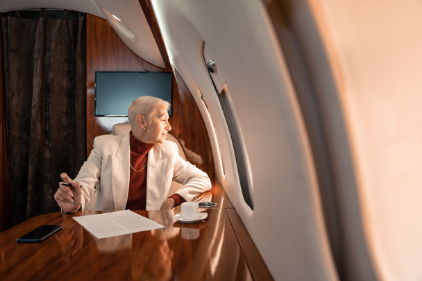 Mosolygó érett üzletasszony néz repülőgép ablak közelében kávé és papír  - Fotó, kép