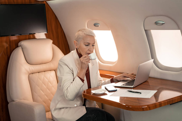 Empresária bebendo café e usando laptop perto de documento no avião  - Foto, Imagem