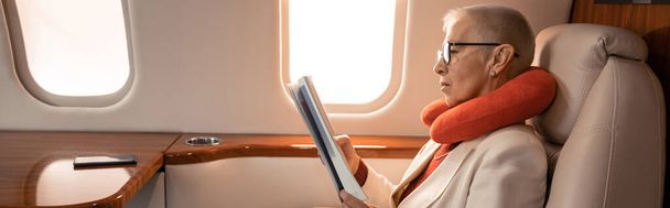Вид сбоку зрелой бизнесвумен в подушке для чтения журнала в частном самолете, баннер  - Фото, изображение