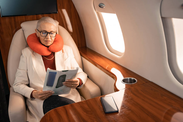 Femme d'affaires dans le magazine de lecture d'oreiller de cou près de smartphone flou dans l'avion  - Photo, image