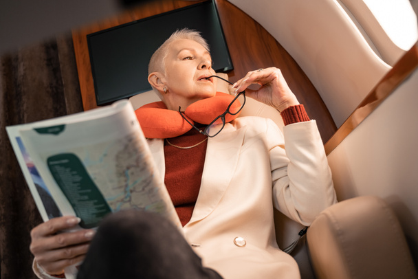Low angle view of businesswoman in neck pillow holding eyeglasses and magazine in private plane  - Photo, Image